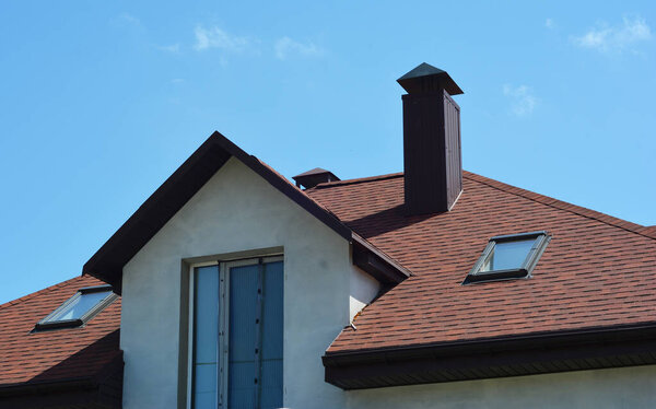 A close-up of an asphalt shingled roofing construction with attic skylights, a chimney, soffit, fascia board and roof gutters installed. 