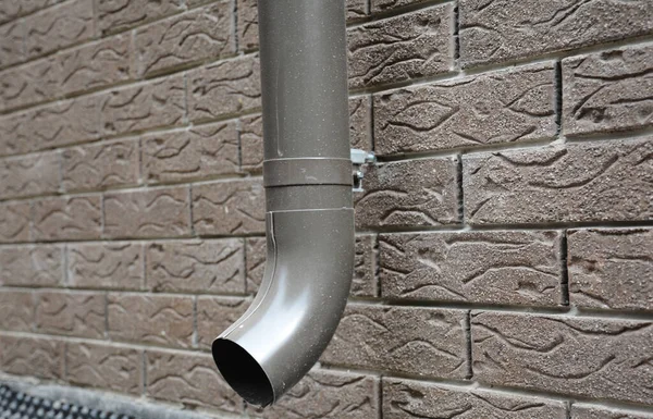 Roof gutter installation: a close-up of a metal rain gutter downspout, downpipe fixed with a bracket, pipe clip to the house foundation, basement tiled with faux stone, brick panels.