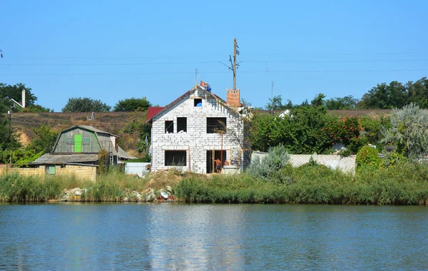 An unfinished brick house under construction on the bank of the river with roofing contractors building a metal roof.