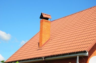 Faded red metal roof tile, rain gutter and chimney against blue sky.