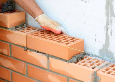 İnşaat işçisi dış cephe oluşturmak için havalanan beton duvarın yanında tuğlalarla çevrili duruyor. İnşaat alanında tuğla dizilimi, çimento uygulaması ve profesyonel tuğla döşeme tekniklerini gösteren duvarcılık çalışmalarının yakın çekimi.