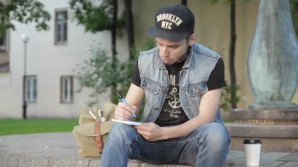 Человек в шапке сидит на гранитной поверхности и рисует что-то синим маркером в блокноте — стоковое видео