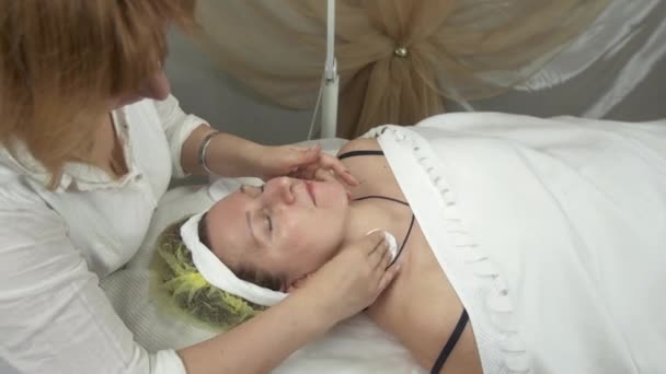 Косметолог витирає обличчя, шию мокрою серветкою для дорослої жінки в салоні краси — стокове відео