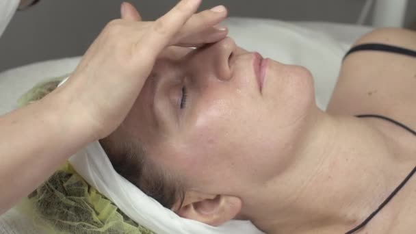 Cosmetologue mettre de l'huile humidifier sur le visage de la femme adulte par les mains dans le salon de beauté . — Video