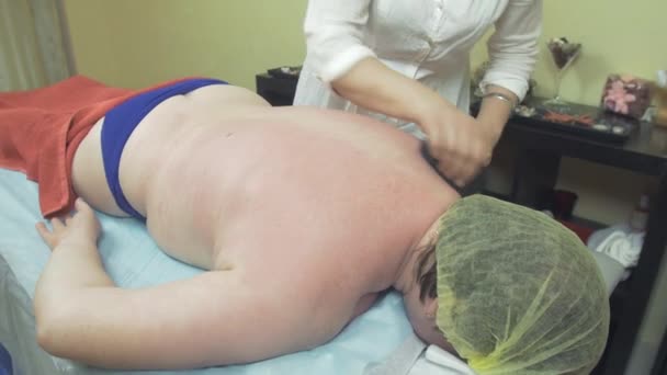 Masseuse make stone therapy massage of left back side to thick woman in saloon. — Stock Video