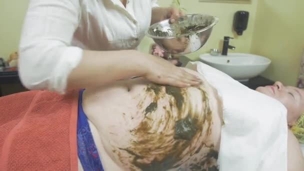 Косметолог поклав суміш морських водоростей на жирний живіт жінки, ліва рука в салоні — стокове відео