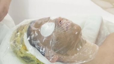 Woman face with green mask, covered by thermofilm in beauty saloon. Warming mask
