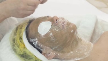 Kozmetolojist salonda yeşil maskeli kadın yüzüne termofilm koydu. Pamuk ped