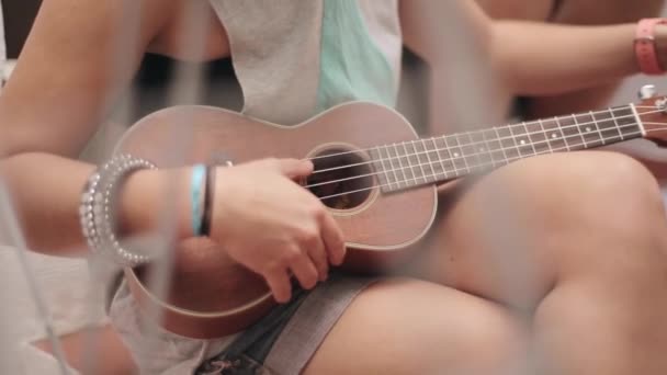 Ragazza seduta in pantaloncini sintonizzare chitarra ukulele marrone sulla strada. Giornata estiva di sole. Musica. Stringhe. Suono — Video Stock