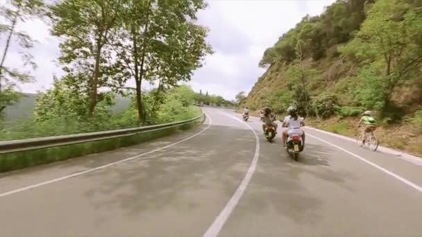 Люди в шоломах їздять на мотоциклі по дорозі в гори, повні зелених дерев. Хвильова рука перехожим — стокове відео