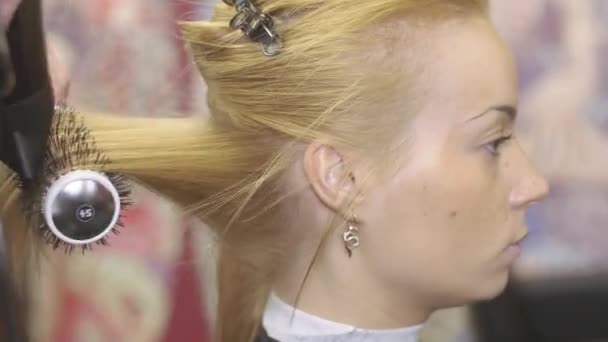 Парикмахер тянет волосы блондинки за расческу и фен в салоне красоты. Создание объемной прически — стоковое видео