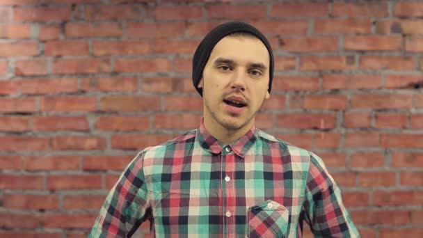 Joven blogger con sombrero negro, camisa a cuadros pronuncia palabras en cámara. pared de ladrillo en el fondo . — Vídeo de stock