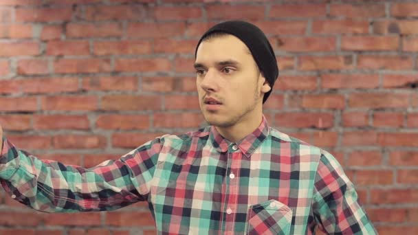 Blogger in black hat, plaid shirt speak words in camera with raised hand. Brick wall on background — Stock Video