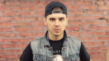 Blogger siyah şapkalı, kot ceket telaffuz kelimeler kamera. Jest. Arka plan üzerinde tuğla duvar.