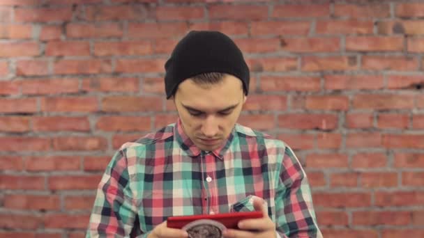 Blogger en sombrero negro, camisa a cuadros toque en la tableta roja delante de la cámara. pared de ladrillo en el fondo . — Vídeo de stock