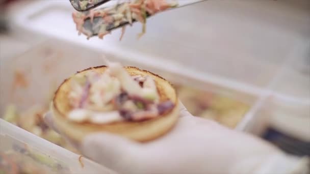 Μαγειρεύετε σε γάντια βάλει τα σαλάτα λαχανικών σε κουλούρι για χάμπουργκερ. Fast food. Καλοκαιρινή μέρα. Ανοιχτό αέρα γεγονότος — Αρχείο Βίντεο