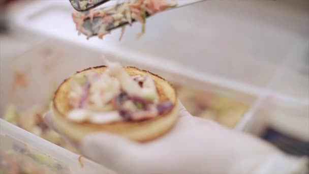 Cocinen en los guantes pongan la ensalada vegetal al bollo para la hamburguesa. Comida rápida. Día de verano. Evento al aire libre — Vídeos de Stock