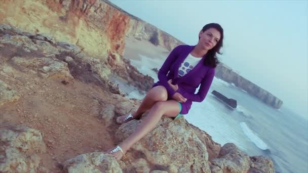 Chica joven morena en chaqueta púrpura, pantalones cortos sentados en la roca en el mar. Noche de verano. Vacaciones. Sonríe — Vídeo de stock