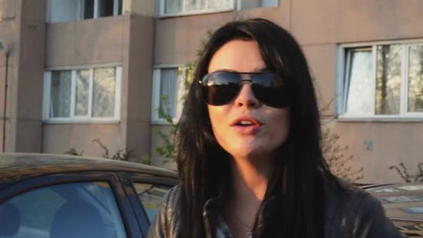 Brunette girl in sunglasses sing in camera at house on street in summer sunny evening. Black jacket — Stock Video