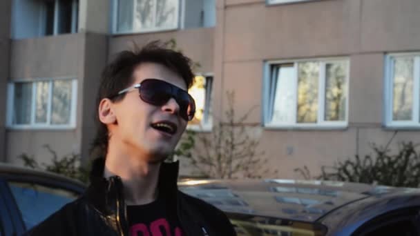 Brunetka człowiek w okulary czytaj rap w aparacie w domu na ulicy w letni wieczór. Czarna kurtka. — Wideo stockowe