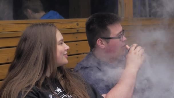 Lány és ember füst elektronikus cigaretta ül a padon. Vapers. Steam. — Stock videók