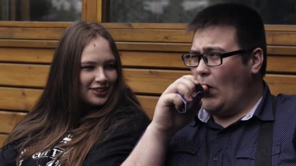 Girl and man in glasses smoke electronic cigarettes on bench. Vapers. Steam. — Stock Video