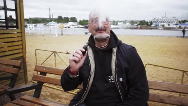 L'uomo anziano fuma sigaretta elettronica sulla spiaggia sul lungomare sorridere in macchina fotografica. Vaper . — Video Stock
