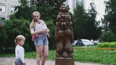 Young mother with baby on hands and son walk on playground with wooden figures