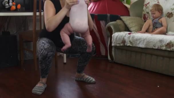 若い母親は、赤ちゃんの手の中でスクワットを作る。息子がソファーに座っていた。アパート — ストック動画