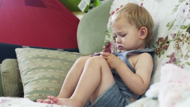 Bedårande liten pojke sitter på soffan i lägenheten knacka i smartphone. — Stockvideo