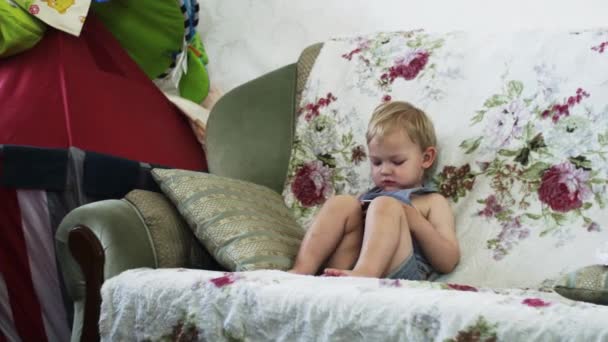 Ξανθιά μικρό αγόρι κάθεται στον καναπέ στο διαμέρισμα βρύσης σε smartphone. Μητέρα με μωρό. — Αρχείο Βίντεο