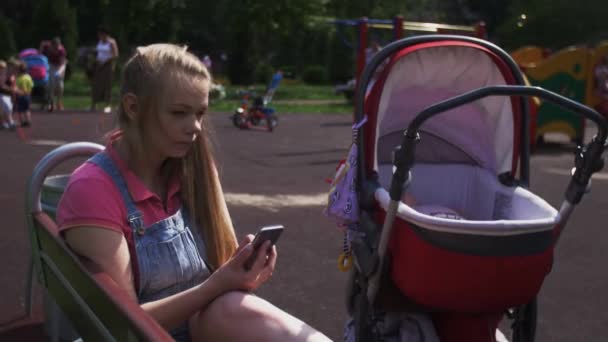 Madre joven sentarse en el banco en el patio de recreo, tocando en el teléfono inteligente. Carruaje bebé . — Vídeos de Stock