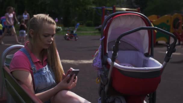 Mother sit on bench on playground tap on smartphone. Red baby carriage. Children — Stock Video