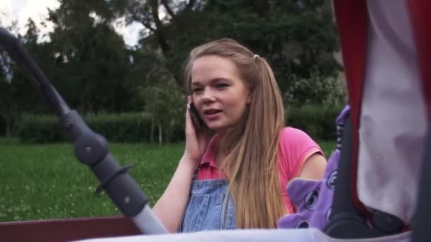 Ung mamma tala med telefonen på bänk. Barnvagn. Sommardag. Leende — Stockvideo