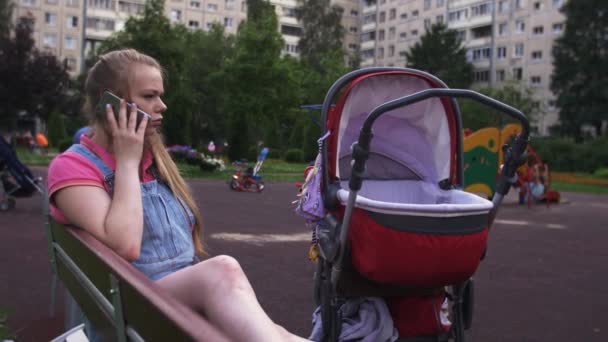 Ung mamma tala med telefon på lekplatsen. Röd barnvagn. Barn. Sommaren — Stockvideo