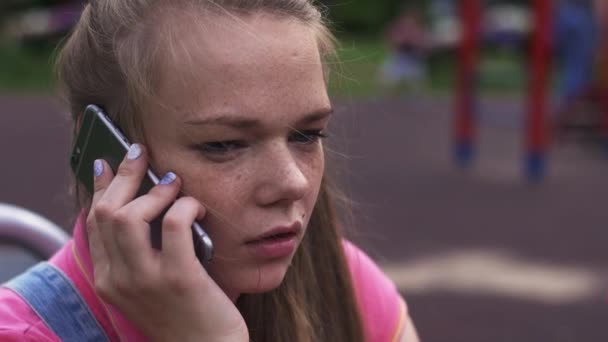 Unga attraktiva flicka med fräknar tala med telefon på lekplatsen. Sommaren park. — Stockvideo