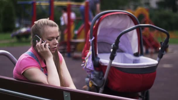 Νεαρή μητέρα μιλούν στο τηλέφωνο σε παιδική χαρά. Κόκκινη άμαξα μωρό. Τα παιδιά. Πάγκος. — Αρχείο Βίντεο