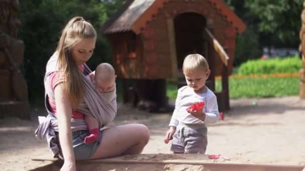 Fiatal anya tartsa baby sling, játszani a homokozóban fia. Játszótér. Süt a nap — Stock videók