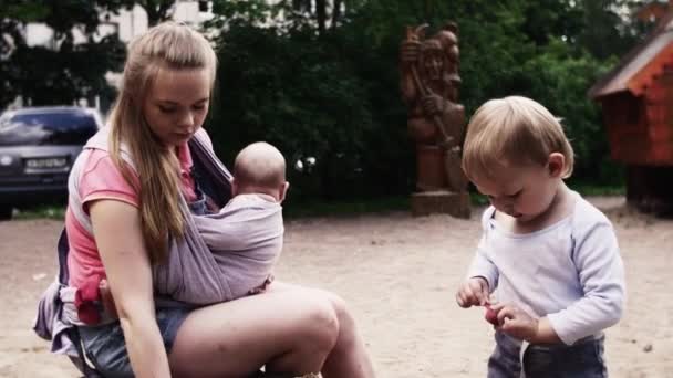 Junge Mutter hält Baby im Tragetuch mit Sohn auf Spielplatz Sommertag. — Stockvideo