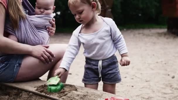 Νεαρή μητέρα κρατήστε το μωρό στη σφεντόνα παιχνίδι με γιο στο sandbox σε παιδική χαρά. Το καλοκαίρι. — Αρχείο Βίντεο
