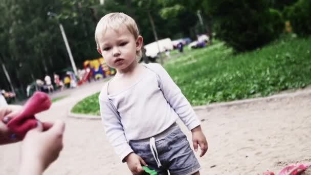 Młoda matka umieścić na skarpety na dziecko w chuście, syn grać w piaskownicy. Letni dzień. — Wideo stockowe