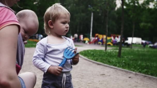 Молода мати тримає милу дитину в слінгу грати з сином у пісочниці. Літо. парку — стокове відео