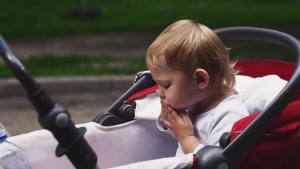 Lilla blonda pojken äta kinder överraskning i barnvagn på gatan. Sommardag — Stockvideo