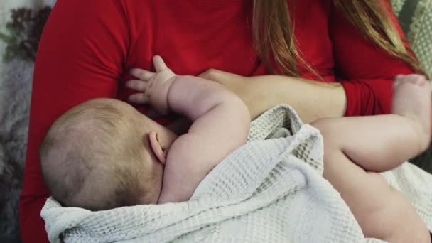 Молода мати в червоній сорочці годує маленького плачучого немовляти на дивані. Догляд за мамою — стокове відео