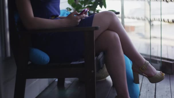 Женщина в синем платье, туфли на высоких каблуках сидят на террасе ресторана со смартфоном — стоковое видео