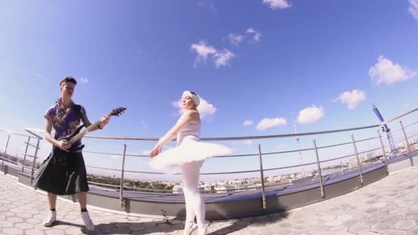 Hombre en falda tocar la guitarra eléctrica en primera línea de mar, cantar en cámara. Baile de niña en traje de bailarina. Loco. — Vídeos de Stock