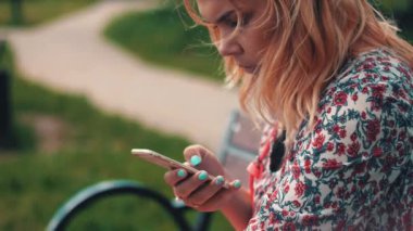 Bankta oturan ve parkta smartphone kullanarak genç kadın. Yaz günü. Gülümseme