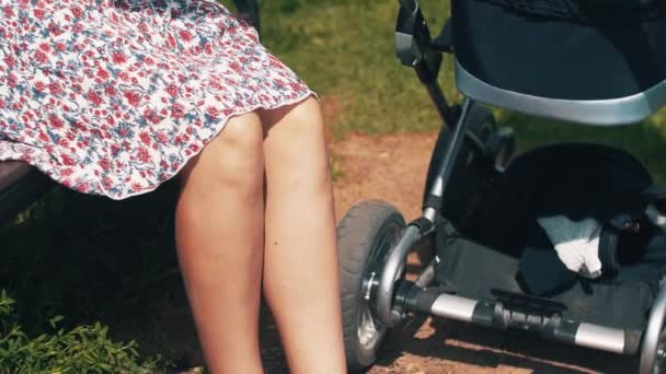 Mãe no vestido de verão balançar a carruagem do bebê sentado no banco. Ensolarado. Maternidade — Vídeo de Stock