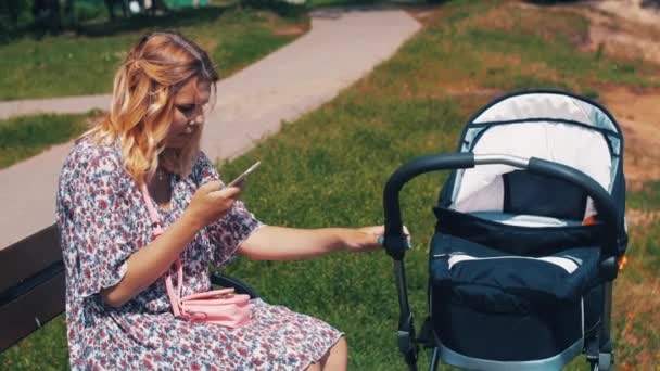 Madre con carro de bebé sentarse en el banco y el uso de teléfono inteligente. Día soleado de verano — Vídeos de Stock