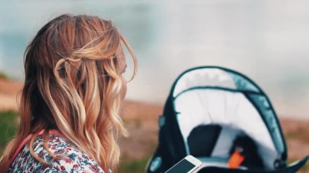 母亲在长凳上晃动婴儿车, 使用智能手机。夏日阳光明媚的日子. — 图库视频影像